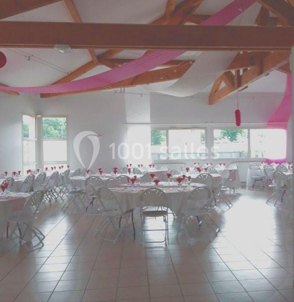 Salle de réception lumineuse avec tables rondes dressées, chaises blanches et décorations roses suspendues.