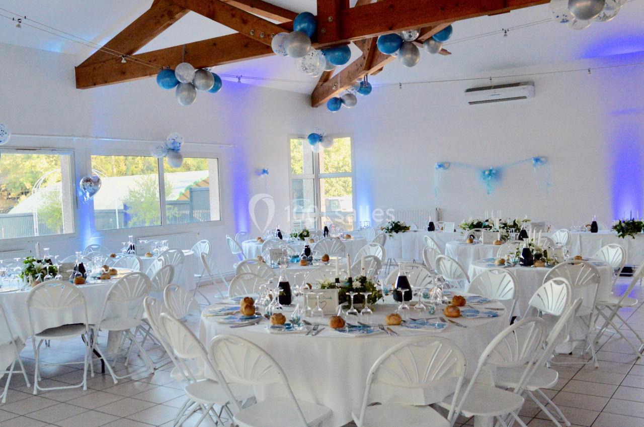 Salle de réception décorée avec des tables rondes dressées, des ballons suspendus et une lumière bleutée.