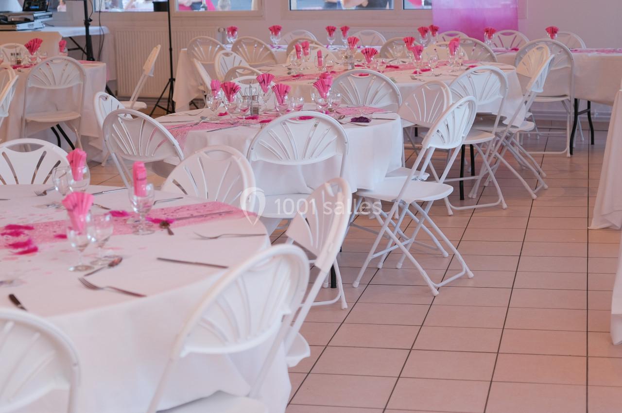 Salle décorée pour un événement, avec des tables rondes dressées, nappes blanches et accents roses.