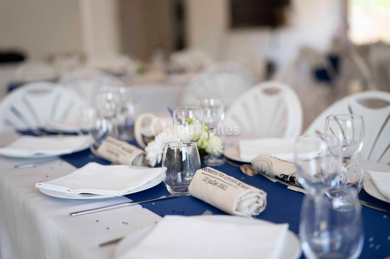 Table dressée avec nappes blanches, chemins de table bleus, serviettes roulées, verres et assiettes dans une salle lumineuse.
