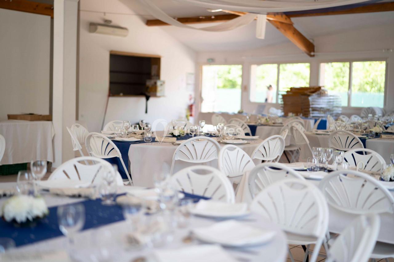 Salle de réception lumineuse avec des tables dressées, nappes bleues et blanches, et chaises en plastique blanc.
