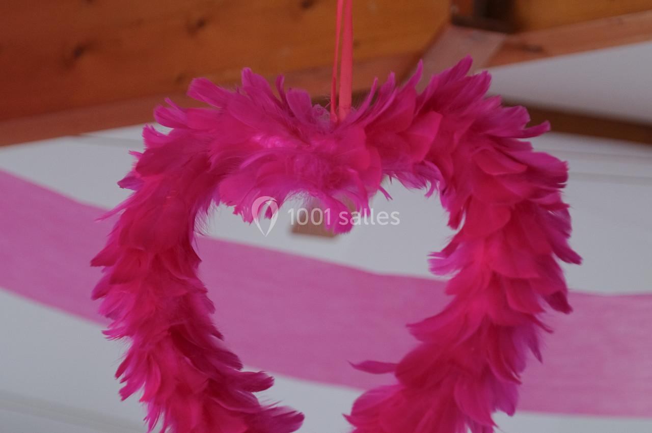 Cœur suspendu en plumes roses, décoré avec un ruban et des éléments roses en arrière-plan.