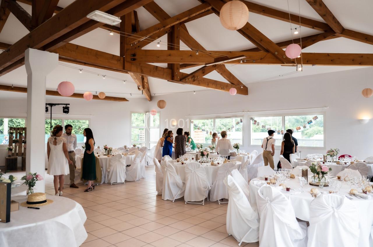 Salle de réception lumineuse décorée pour un événement, avec des tables dressées et des invités en discussion.
