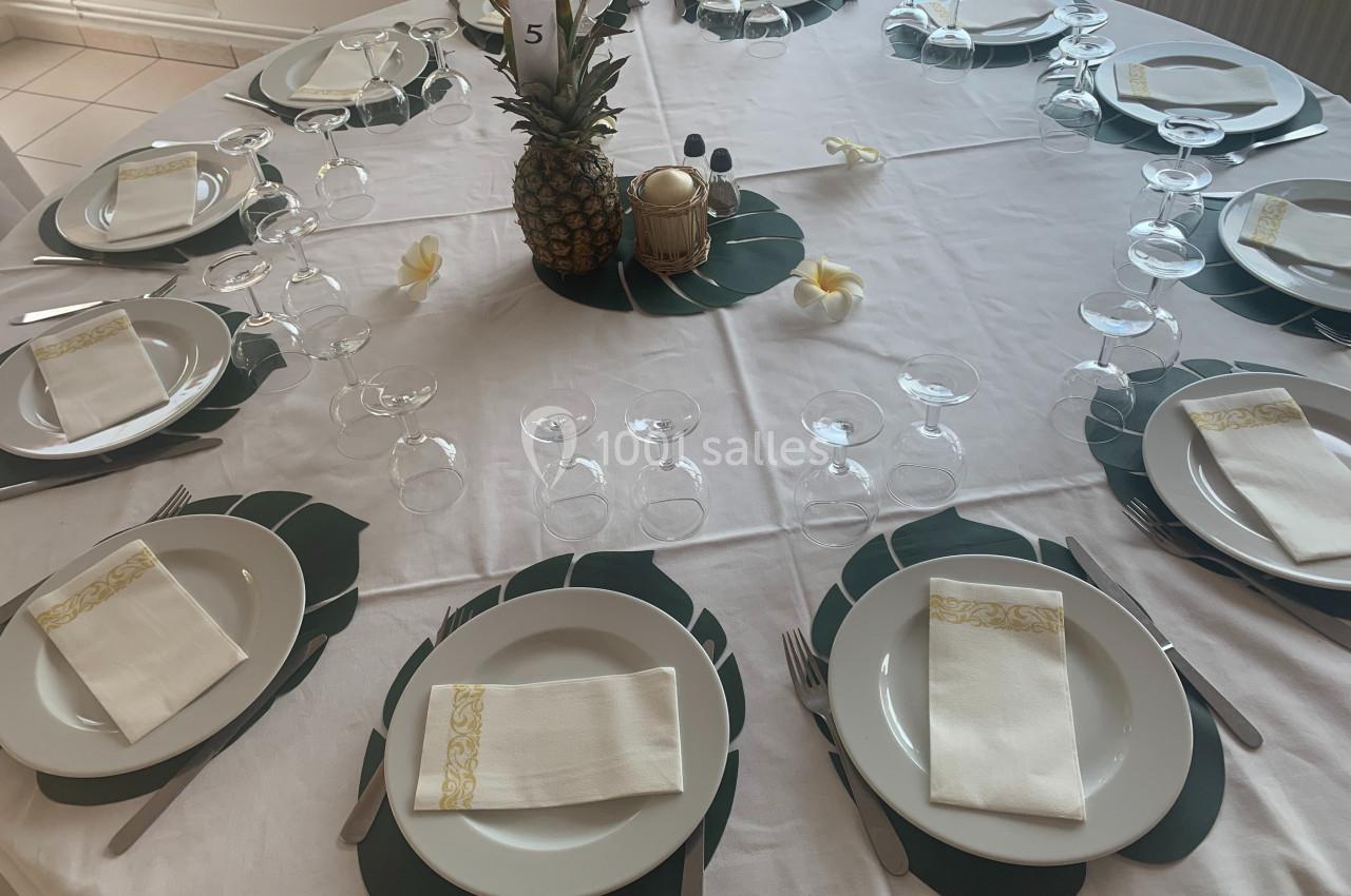 Table ronde dressée avec nappes blanches, assiettes, serviettes pliées, verres et décoration centrale avec un ananas.