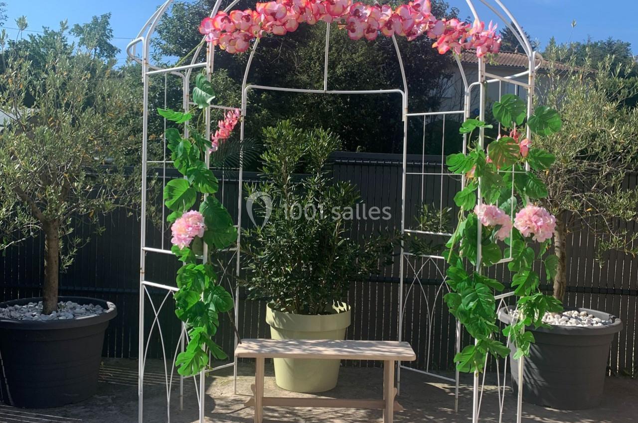 Structure métallique blanche décorée de fleurs roses et feuillages, avec un banc en bois devant un pot d'olivier.