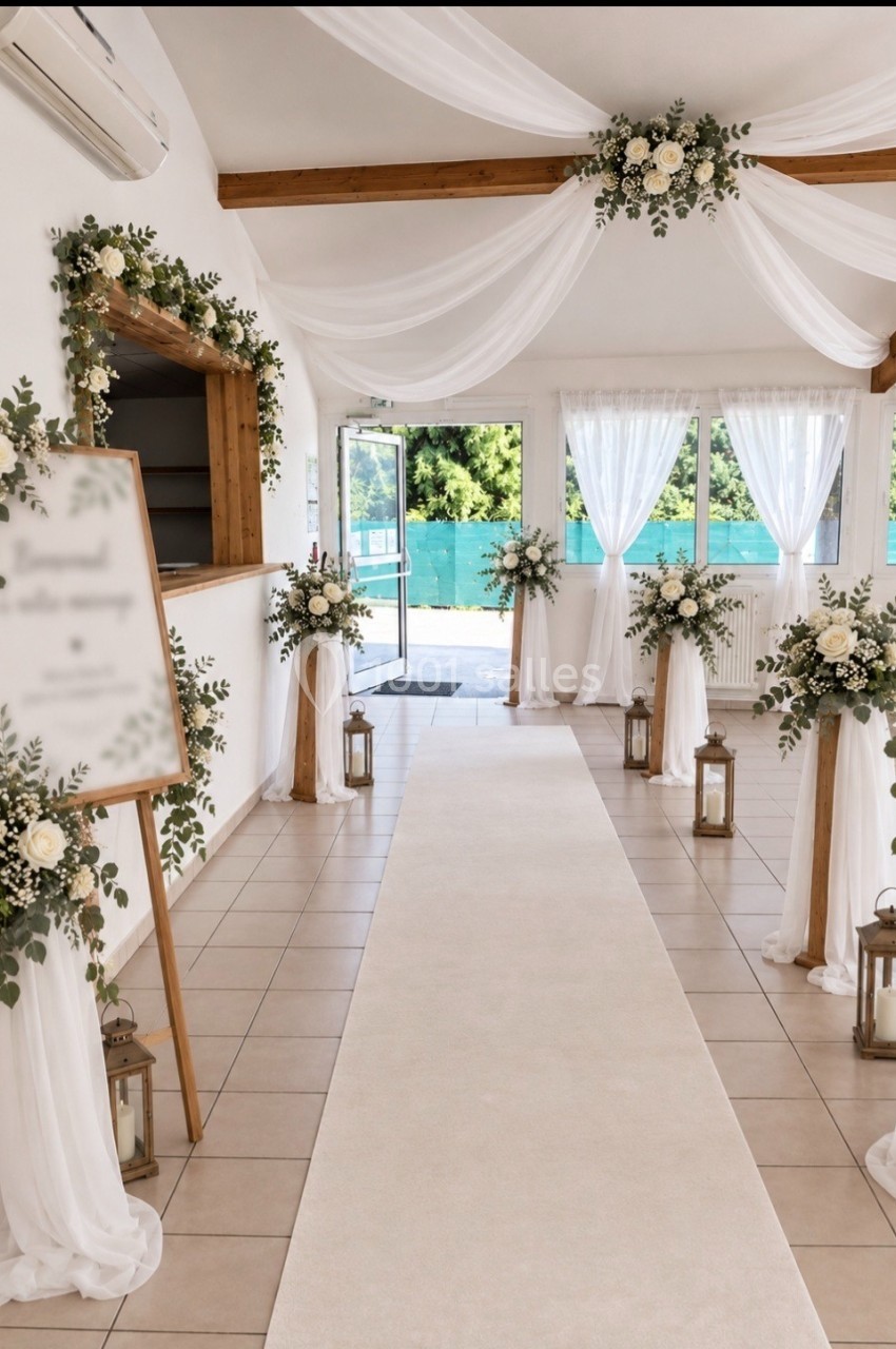 Salle de réception décorée pour un mariage avec allée centrale, fleurs blanches, voilages et lanternes.