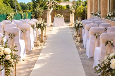 Arche de mariage décorée de fleurs blanches et vertes, avec deux chaises pliantes blanches devant, en extérieur ensoleillé.