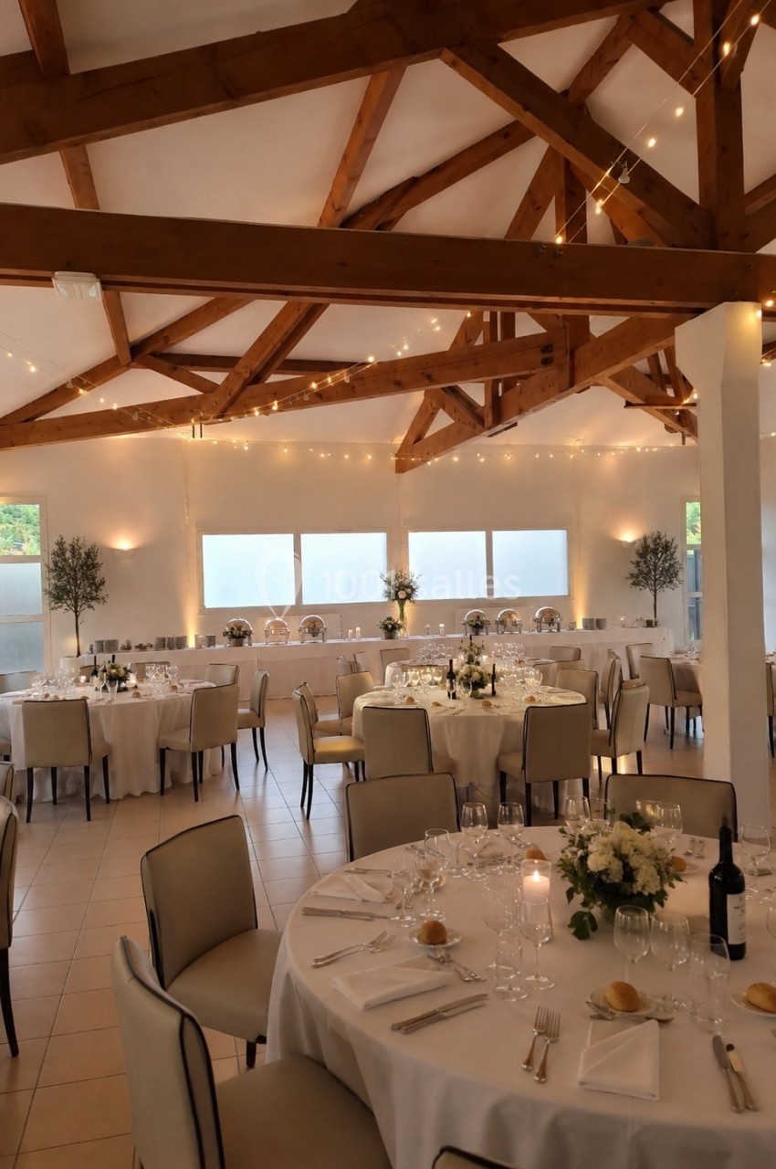 Salle de réception lumineuse avec tables rondes dressées, décorée de guirlandes lumineuses et fleurs blanches.