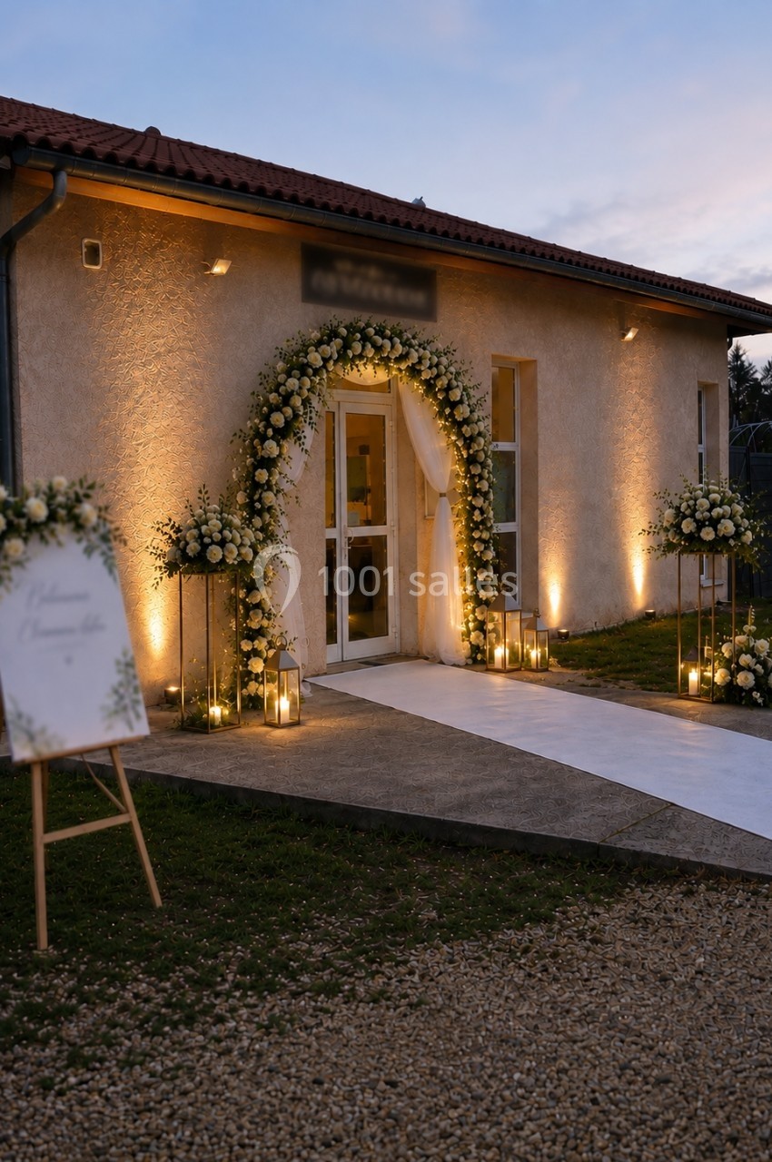 Entrée d'un bâtiment décorée pour un mariage avec arche florale, tapis blanc et lanternes lumineuses.