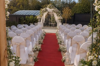 Arche de mariage décorée de fleurs blanches et vertes, avec deux chaises pliantes blanches devant, en extérieur ensoleillé.