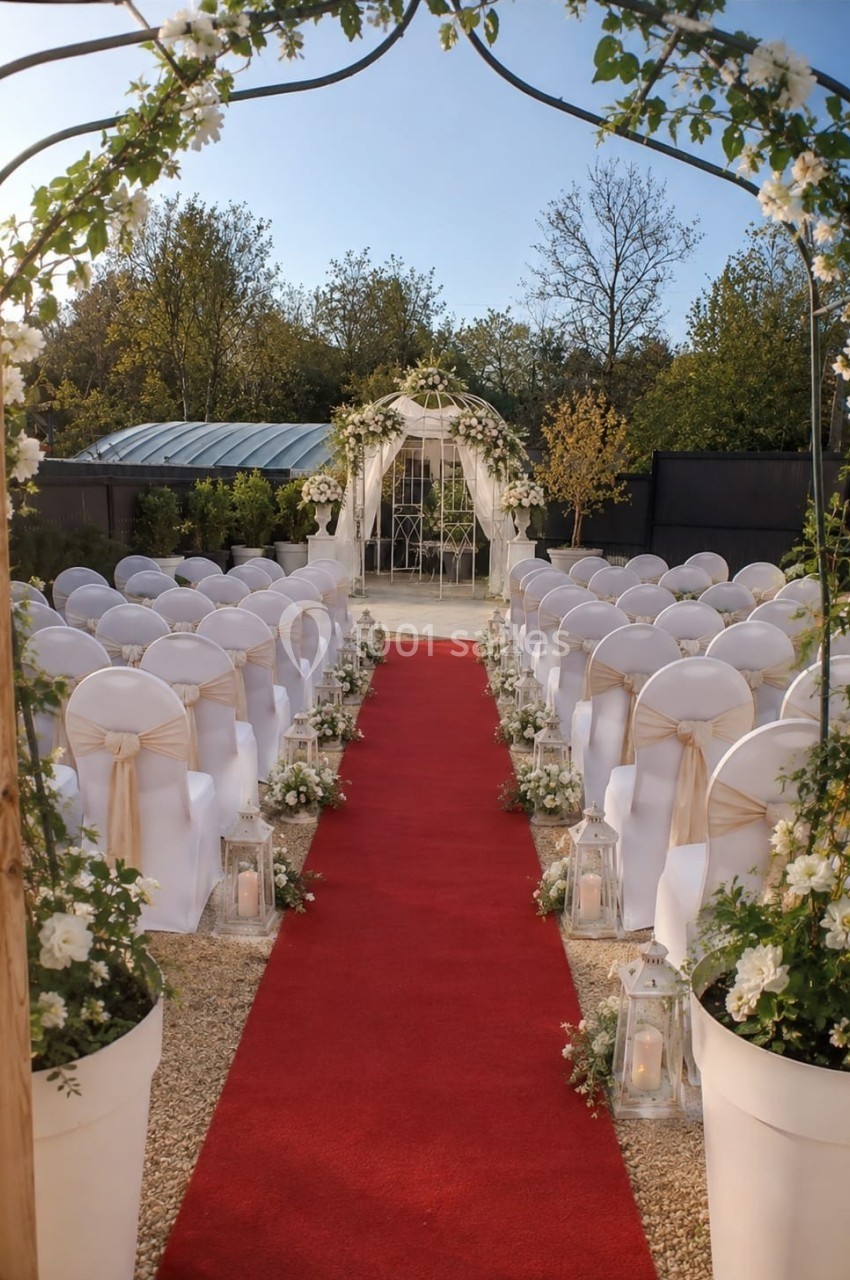 Allée centrale avec tapis rouge menant à une arche fleurie, entourée de chaises décorées pour une cérémonie en extérieur.