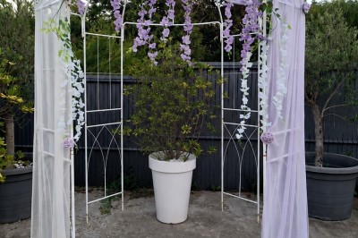 Arche de mariage décorée de fleurs blanches et vertes, avec deux chaises pliantes blanches devant, en extérieur ensoleillé.