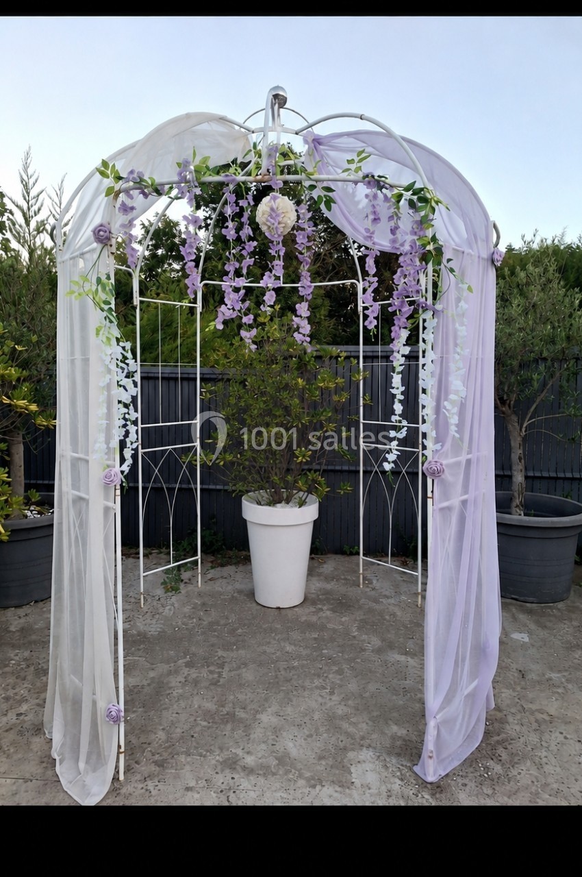 Tonnelle blanche décorée de voiles et de fleurs violettes, placée sur une terrasse avec des plantes en pots.