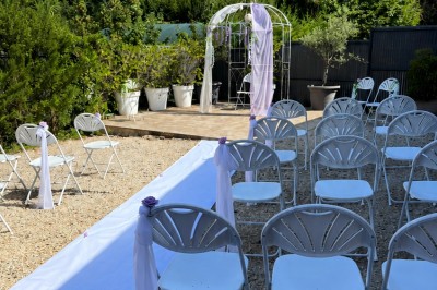 Arche de mariage décorée de fleurs blanches et vertes, avec deux chaises pliantes blanches devant, en extérieur ensoleillé.