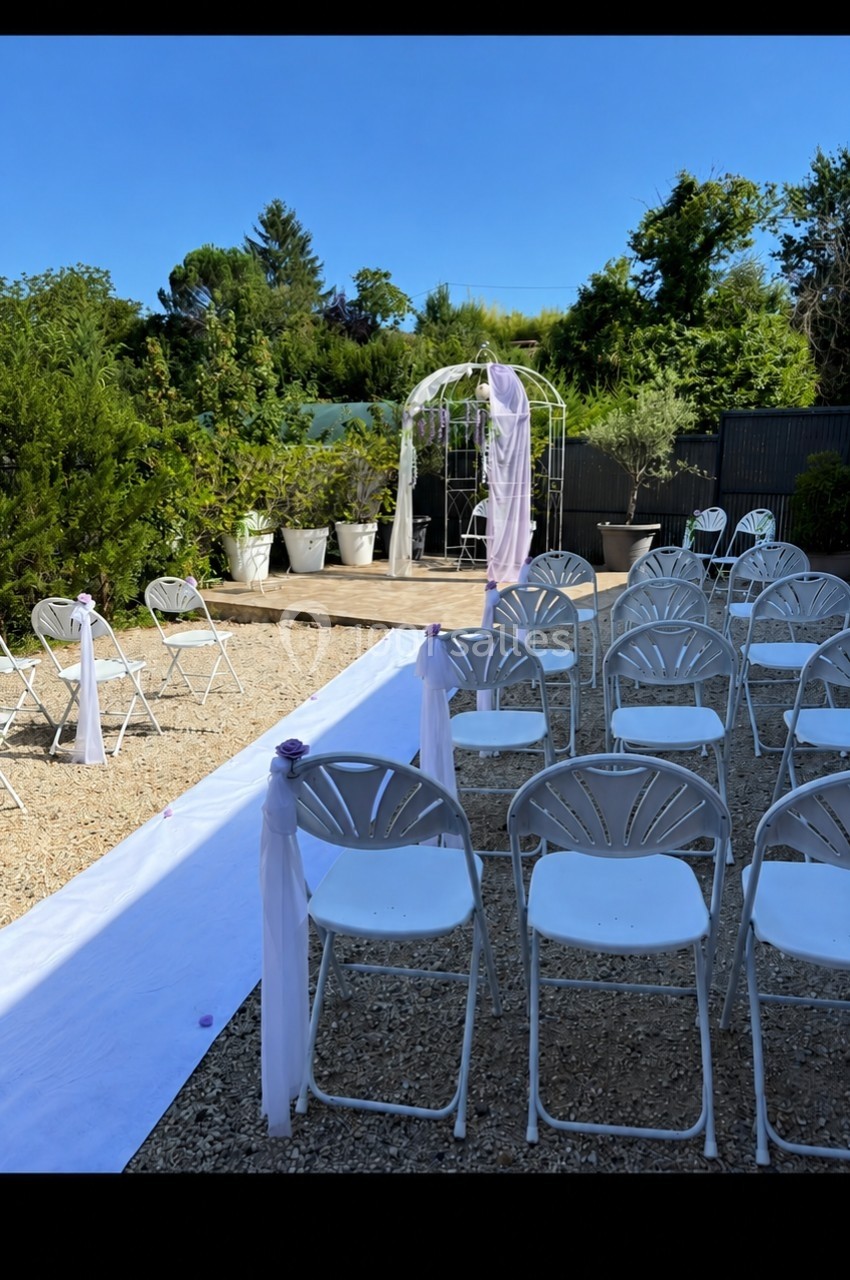 Chaises blanches alignées devant une arche décorée pour une cérémonie en extérieur, sur un sol en gravier.
