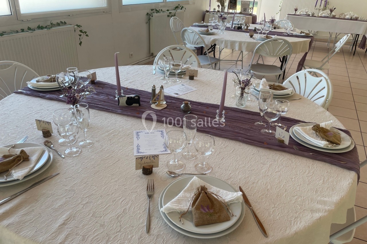 Table décorée pour un repas, avec nappes blanches, chemins de table violets, vaisselle et petits bouquets.