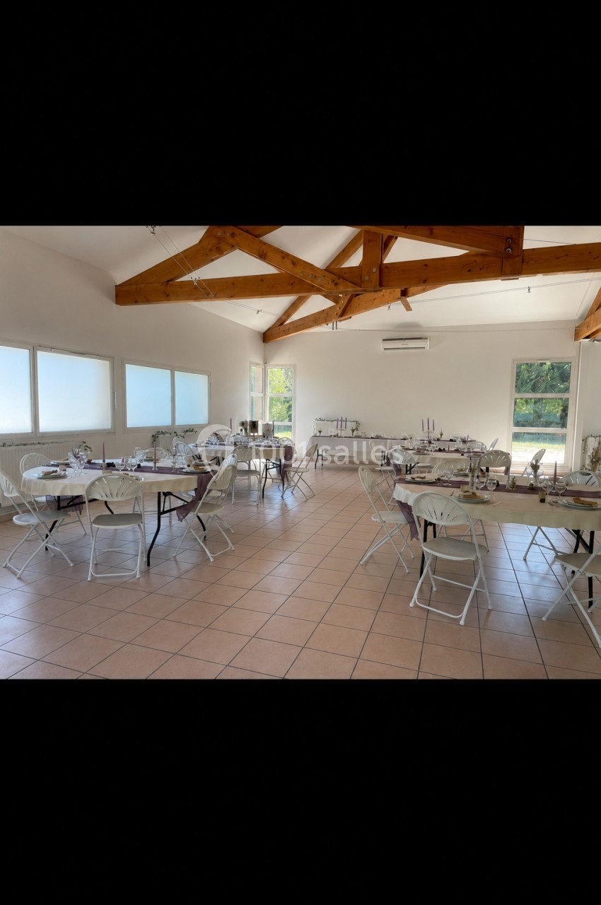 Salle lumineuse avec tables rondes dressées, chaises pliantes blanches et poutres apparentes au plafond.