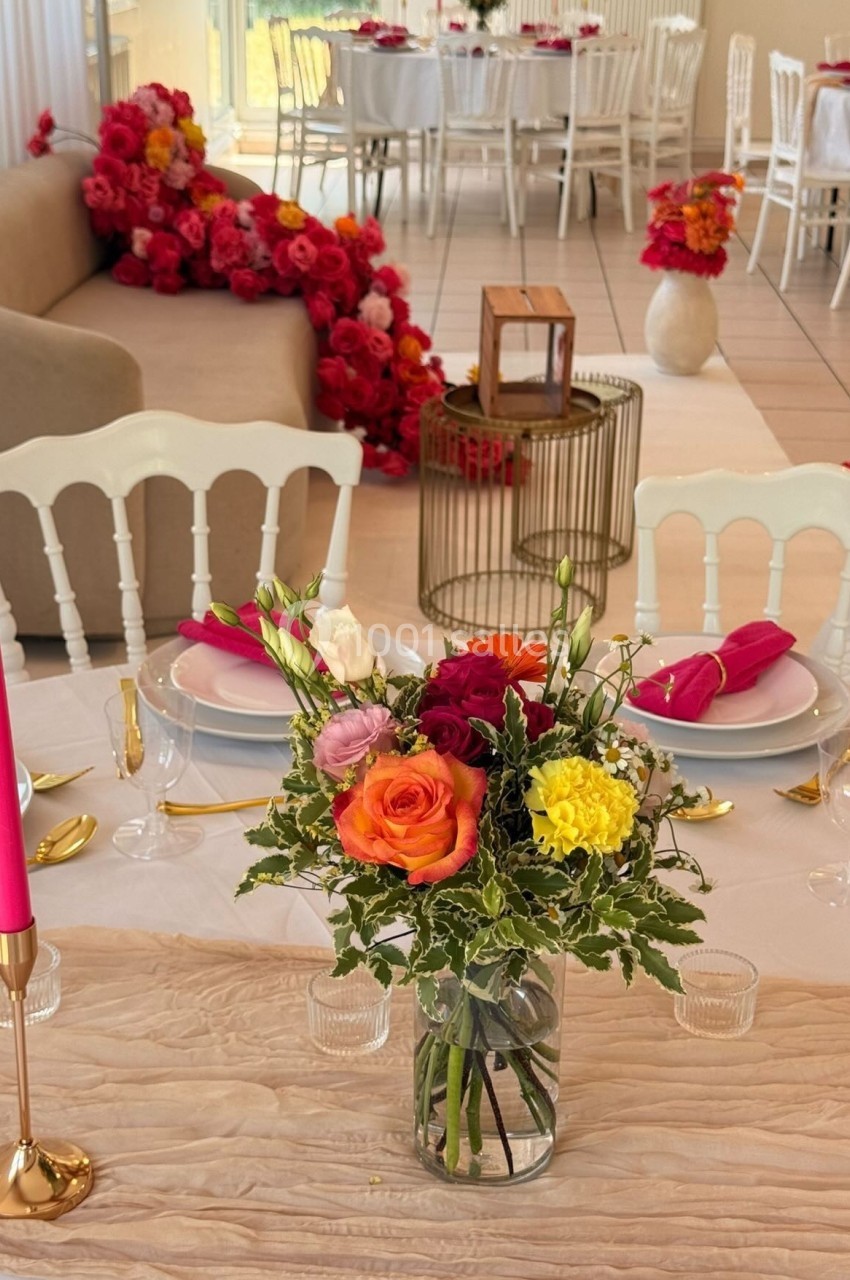 Table décorée avec un bouquet de fleurs colorées, bougie rose et vaisselle élégante, dans une salle ornée de roses rouges.