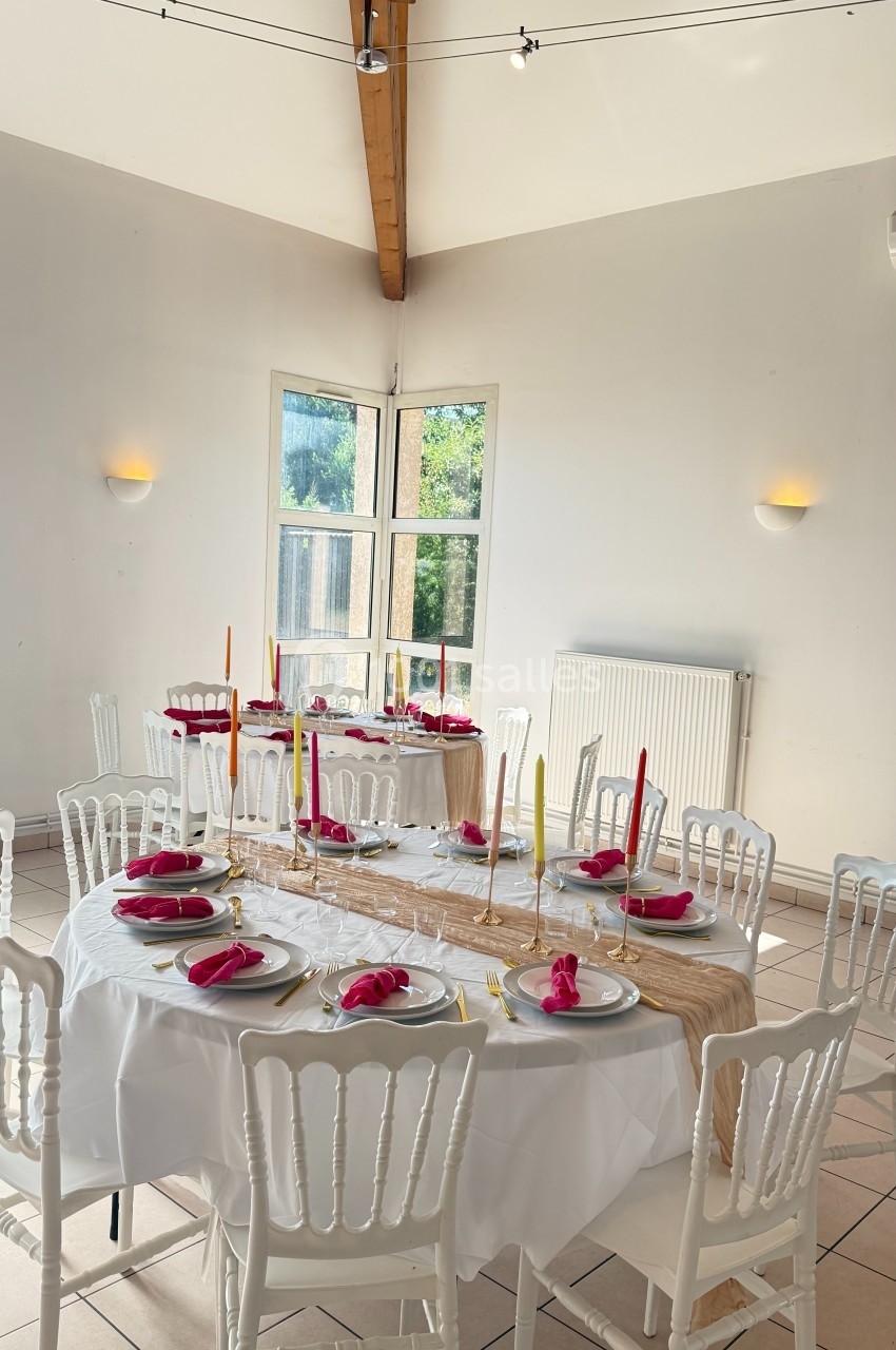 Salle lumineuse avec tables décorées de nappes blanches, bougies colorées et serviettes rouges, prête pour un repas.