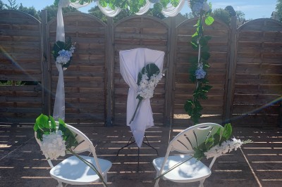Arche de mariage décorée de fleurs blanches et vertes, avec deux chaises pliantes blanches devant, en extérieur ensoleillé.