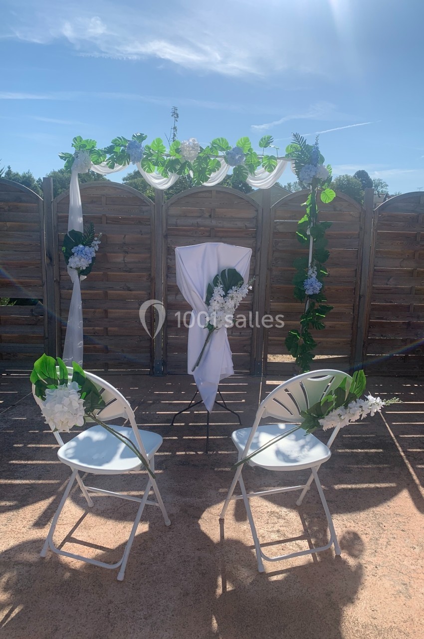 Arche de mariage décorée de fleurs blanches et vertes, avec deux chaises pliantes blanches devant, en extérieur ensoleillé.