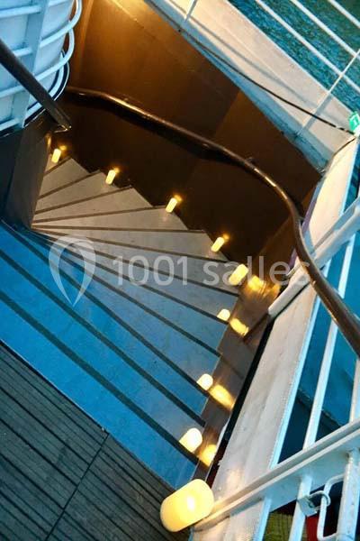 Escalier extérieur en colimaçon éclairé par des lumières au sol, avec une rampe en bois et des marches métalliques.