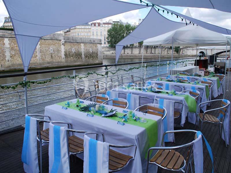 Tables dressées avec nappes blanches, vertes et bleues sur une terrasse en bord de rivière, sous des voiles d'ombrage.