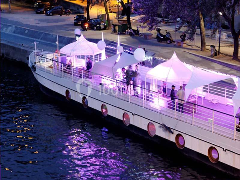 Bateau amarré sur un quai, éclairé par des lumières violettes, avec des tentes blanches et des personnes à bord.