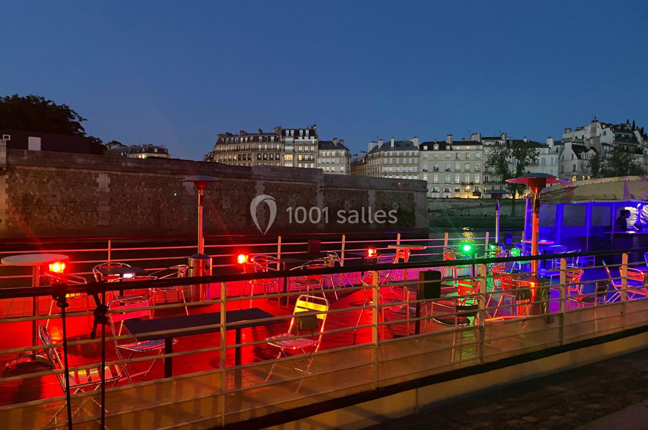 Terrasse de péniche illuminée par des lumières colorées, avec des tables et chaises vides, au bord d'un quai en soirée.