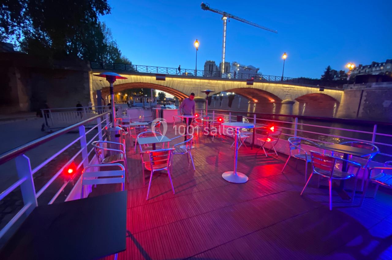 Terrasse extérieure éclairée en soirée avec des tables vides, vue sur un pont illuminé et une grue en arrière-plan.