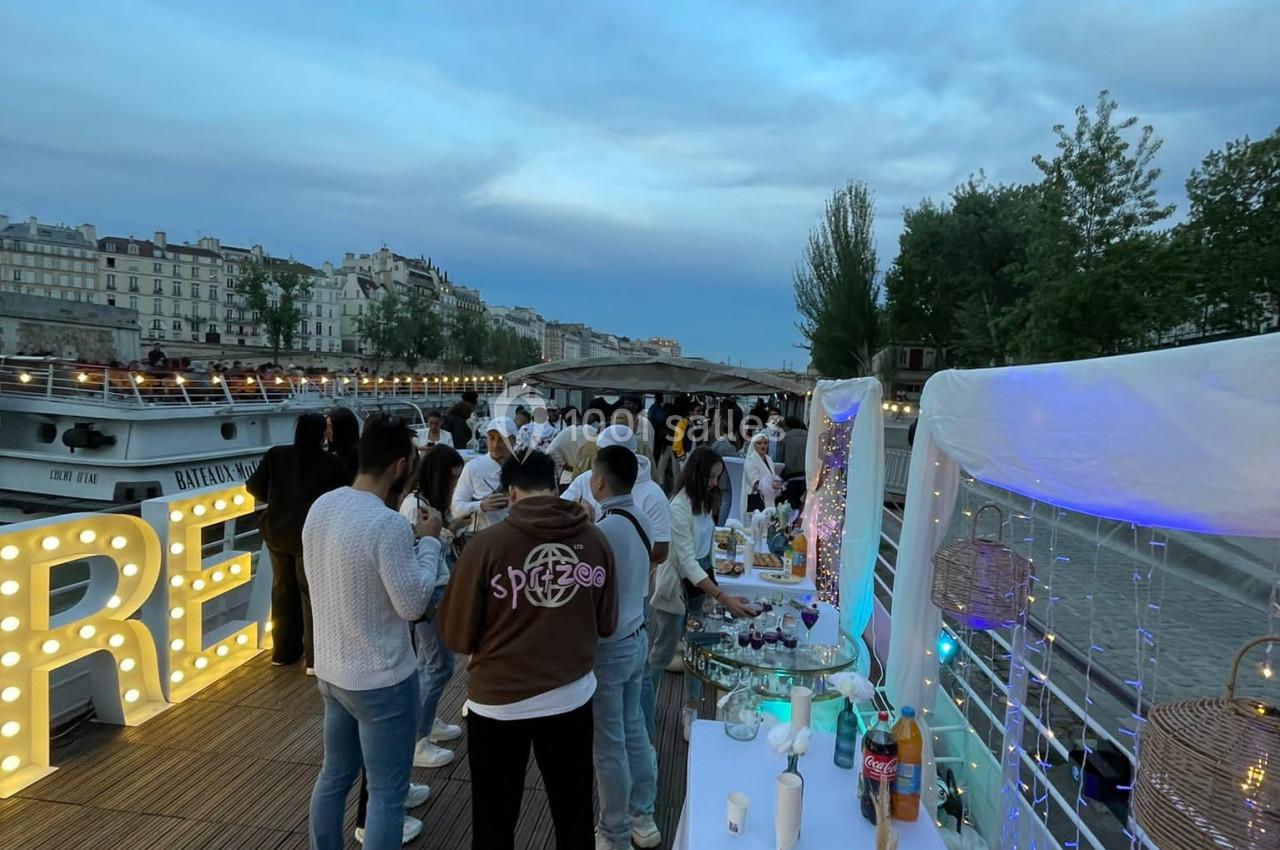 Groupe de personnes rassemblées sur une péniche au bord de la Seine, avec une table de buffet et décorations lumineuses.