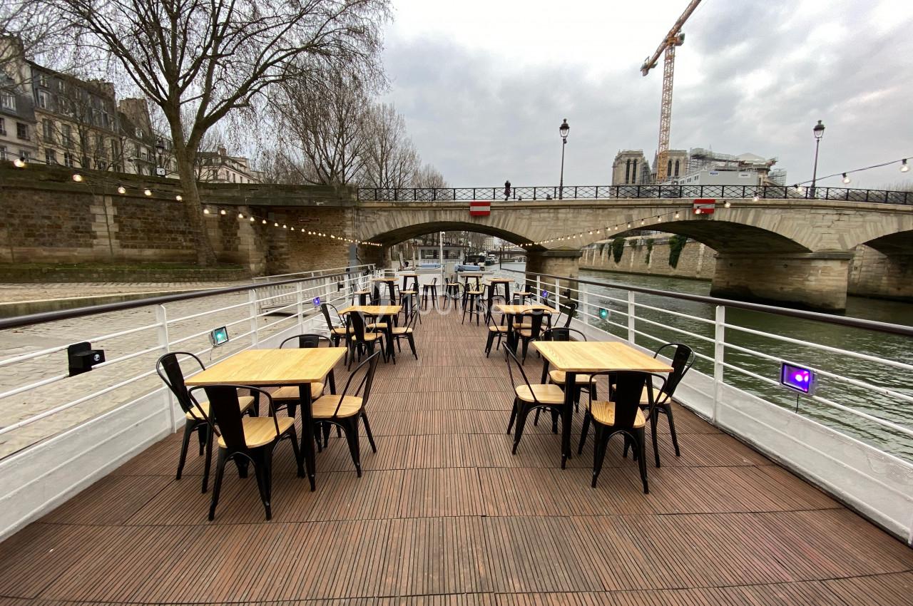 Terrasse en bois avec tables et chaises sur une péniche, vue sur un pont et des bâtiments en bord de Seine.