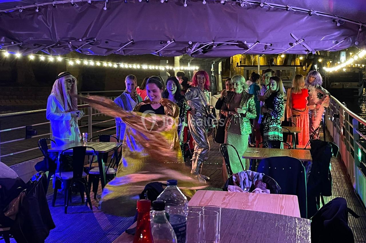 Personnes déguisées dansant et posant sur le pont éclairé d’un bateau, avec des tables et chaises en arrière-plan.