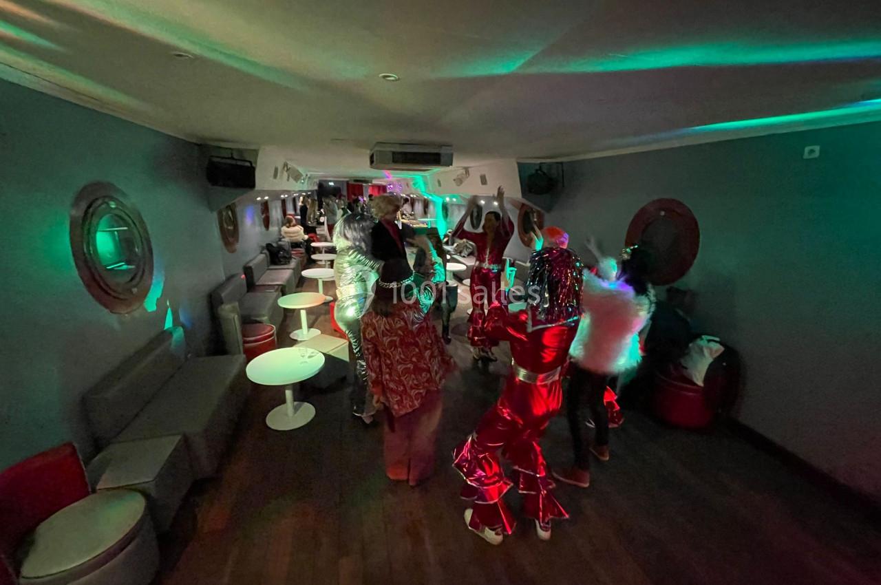 Personnes en costumes colorés dans une salle faiblement éclairée avec tables, banquettes et lumières tamisées.