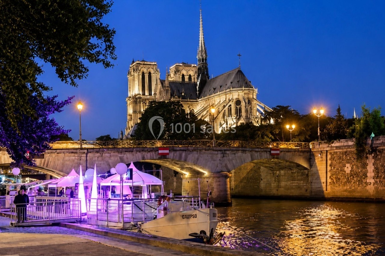 Vue nocturne de la cathédrale Notre-Dame de Paris éclairée, avec un bateau amarré sur la Seine au premier plan.