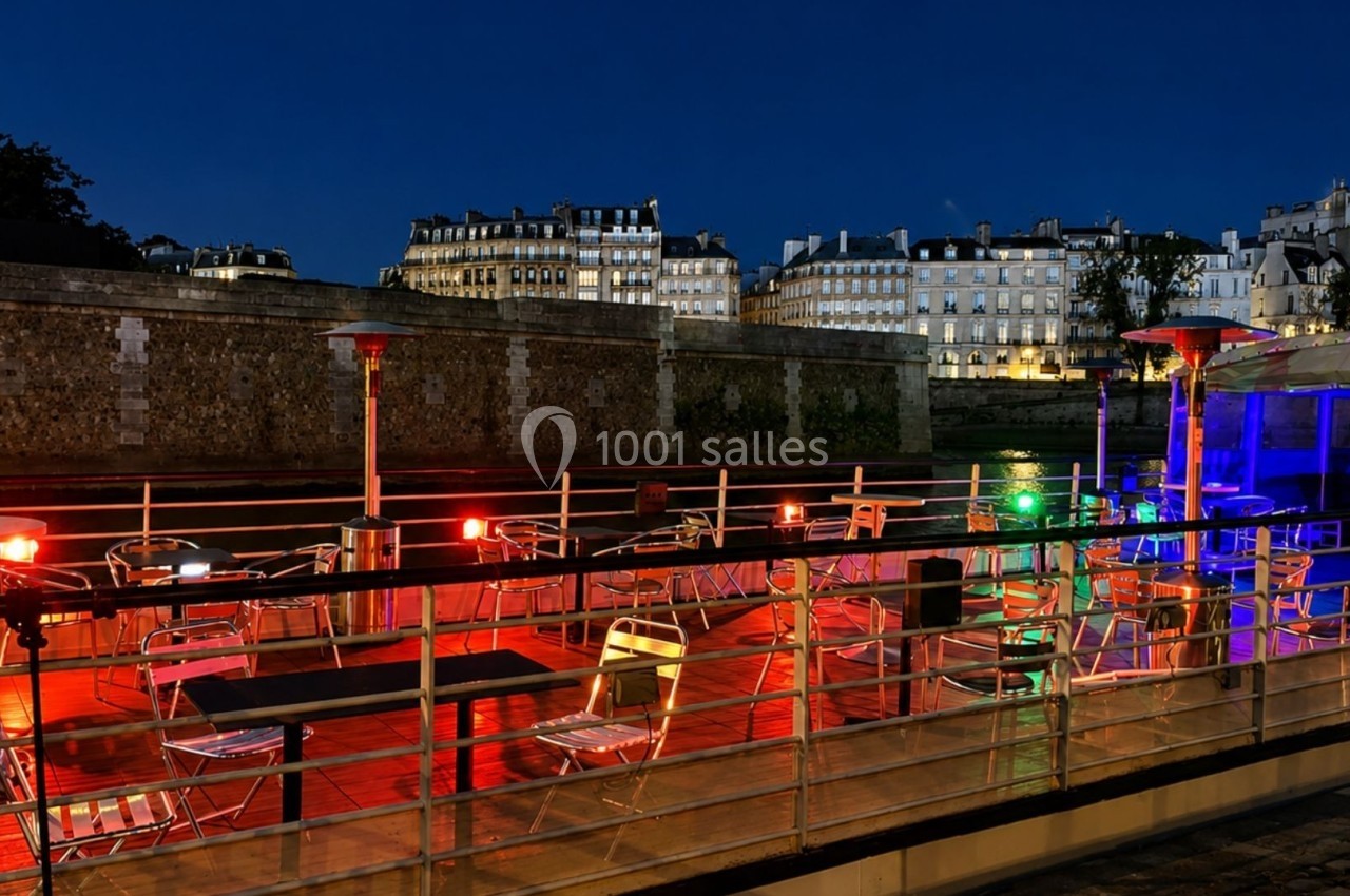 Terrasse éclairée avec des chaises et tables vides sur une péniche, vue nocturne sur des bâtiments historiques en arrière…