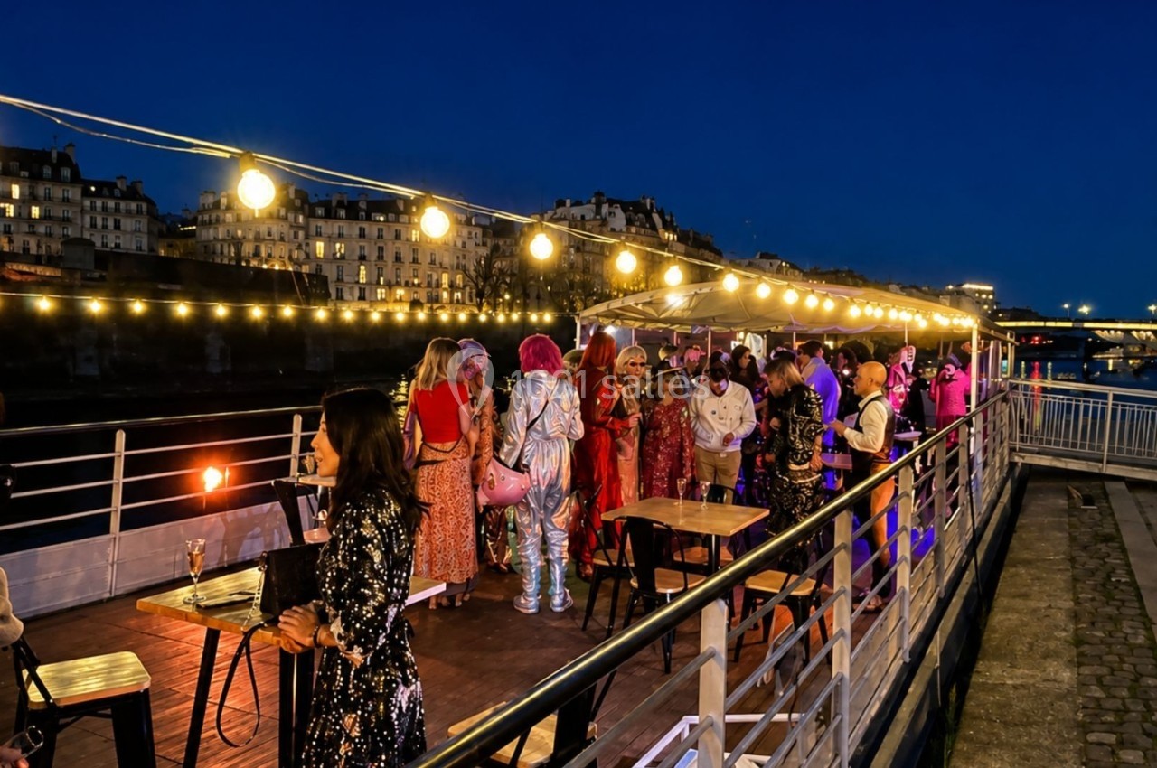 Personnes discutant et célébrant sur une péniche illuminée, amarrée le long d'une rivière en soirée.