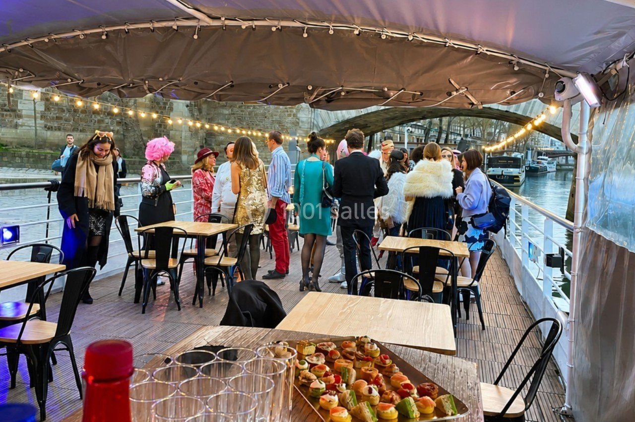 Groupe de personnes en tenue festive rassemblées sur une péniche décorée, avec des tables et des amuse-bouches au premier…