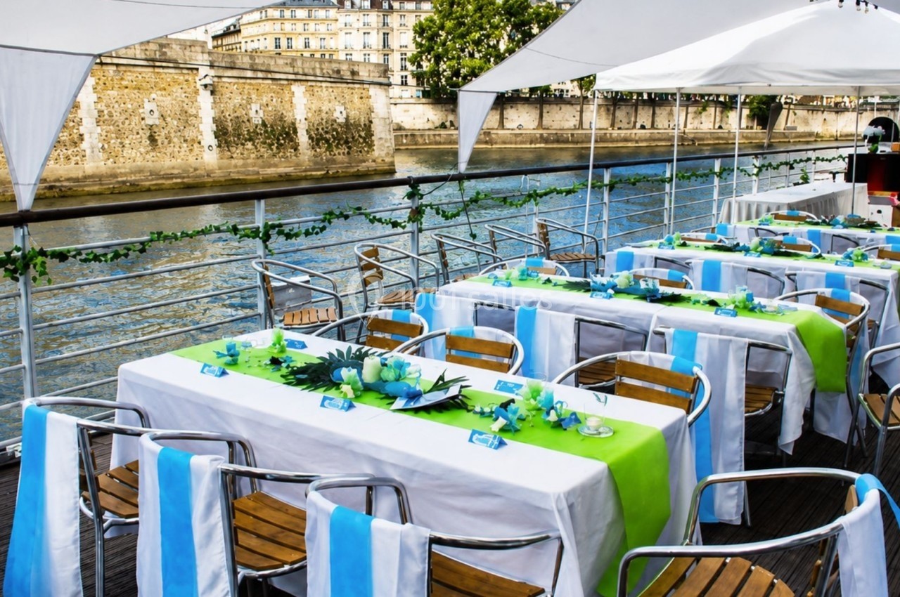 Tables décorées avec nappes vertes et blanches sur une péniche, avec vue sur les quais d'une rivière en arrière-plan.