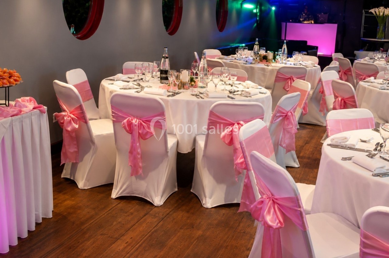 Salle de réception décorée avec des tables rondes, nappes blanches et chaises ornées de nœuds roses.