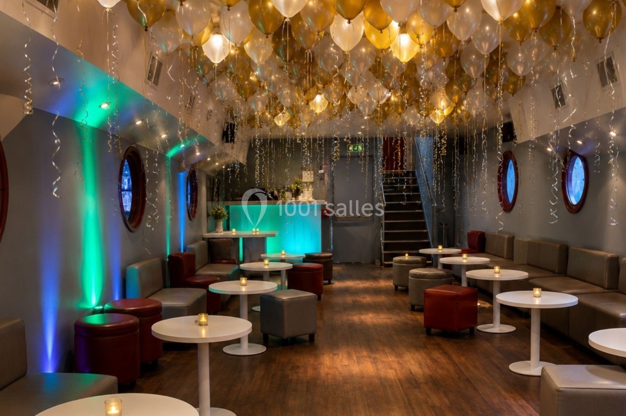 Salle décorée avec des ballons dorés et blancs au plafond, tables basses et éclairage tamisé près d'un bar illuminé.