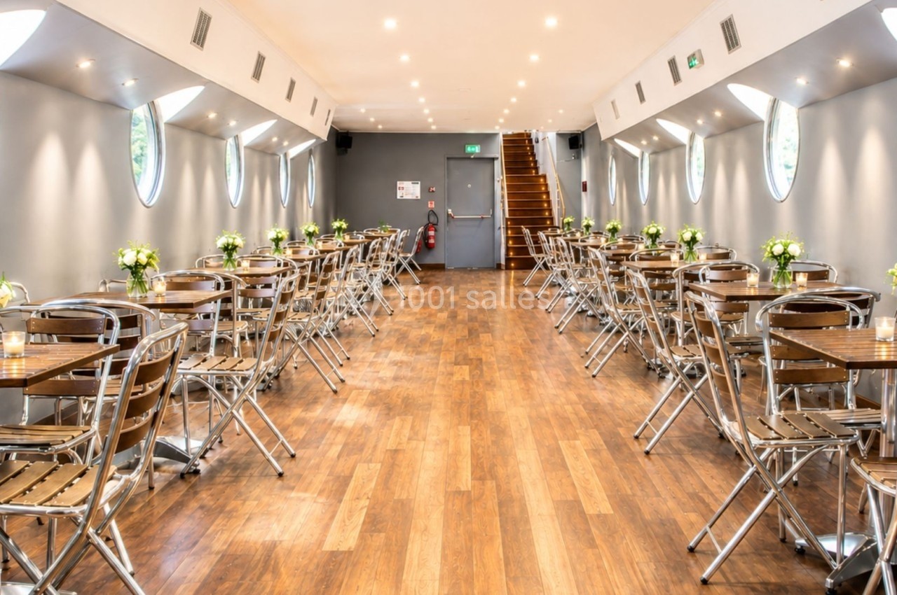 Salle lumineuse avec parquet, tables et chaises en métal, décorée de bouquets, et fenêtres rondes sur les murs.