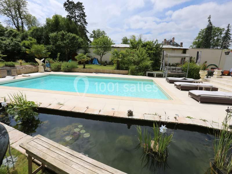 Piscine extérieure entourée de végétation, avec un bassin ornemental au premier plan et des transats sur la terrasse.