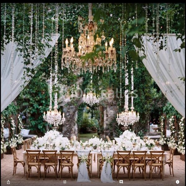 Table élégamment dressée sous des lustres et des guirlandes fleuries, entourée de verdure dans un cadre extérieur.
