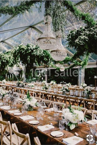 Salle de réception décorée avec des tables en bois, des fleurs blanches, des lustres en cristal et des guirlandes végétales.