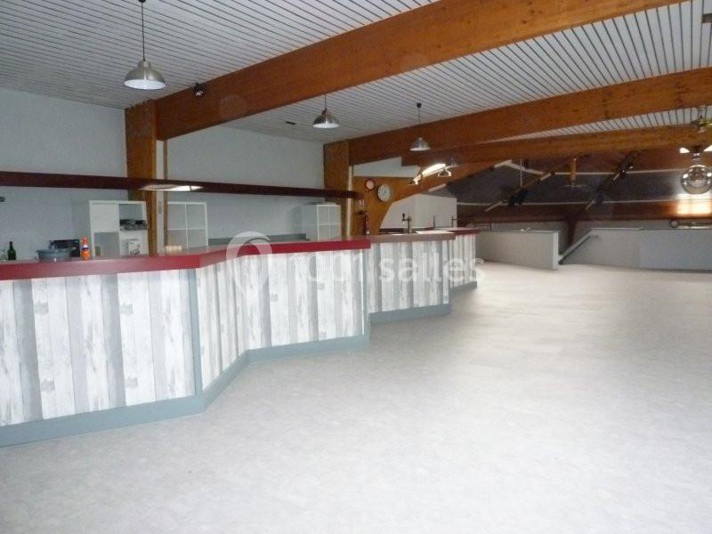 Salle vide avec un long comptoir en bois clair et rouge, plafond à poutres apparentes et sol clair.