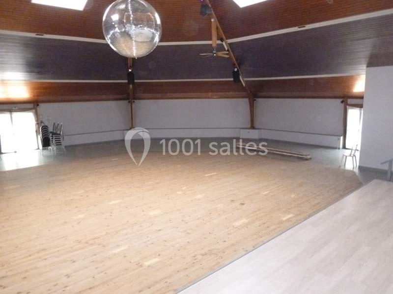 Salle vide avec parquet en bois clair, plafond en pente, grande boule disco suspendue et chaises empilées sur le côté.