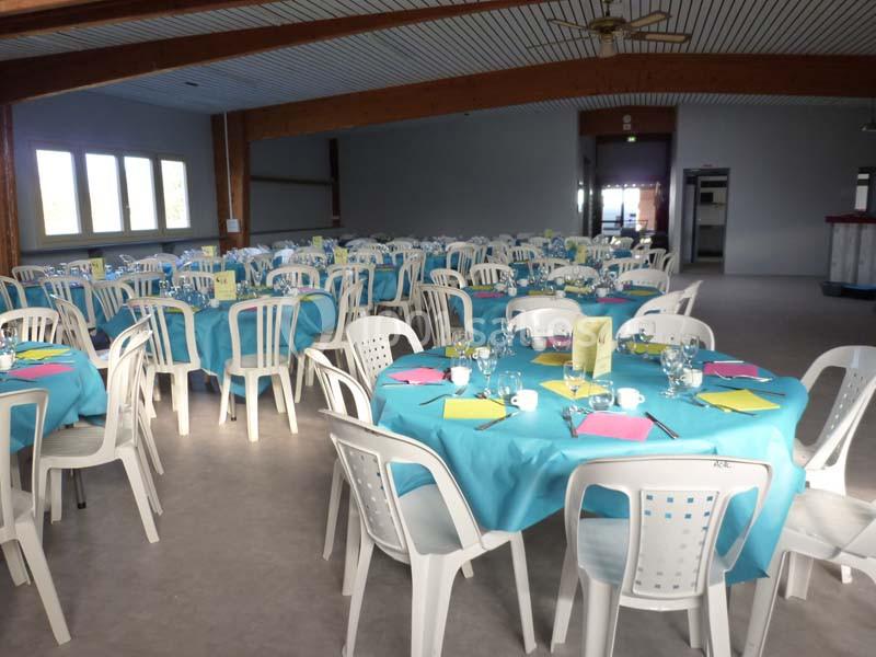 Salle de réception avec des tables rondes dressées de nappes bleues, entourées de chaises blanches en plastique.
