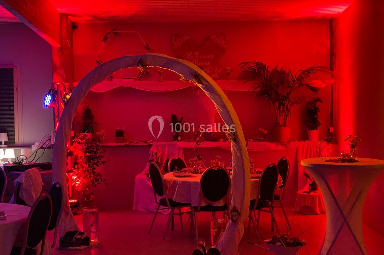 Salle décorée pour un événement, éclairée par une lumière rouge avec tables rondes, plantes et une arche ornée.