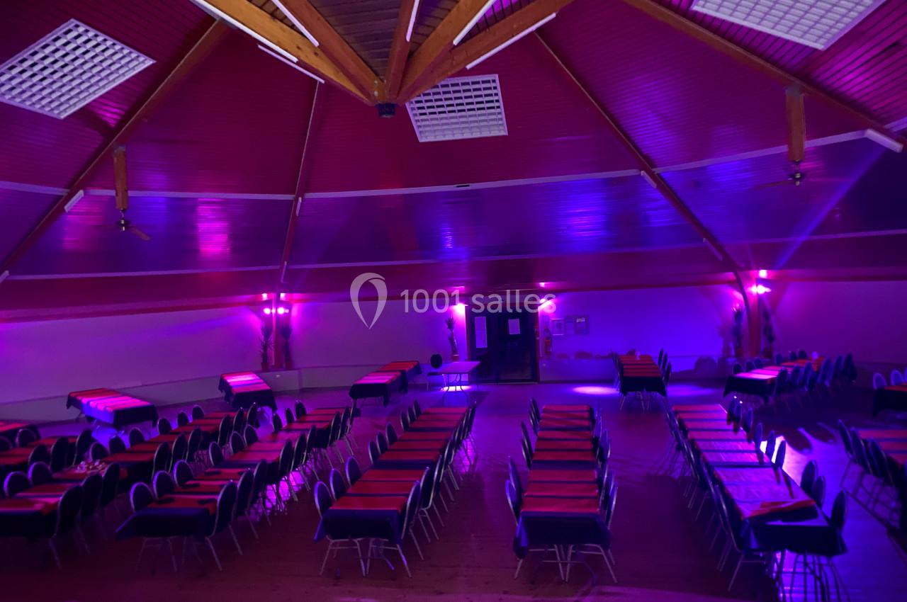Salle de réception avec tables alignées, éclairage tamisé violet et plafond en bois avec poutres apparentes.